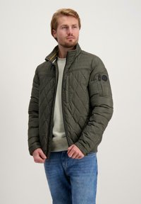 Gevoerde olijfgroene jas met een staande kraag, ritssluiting en een zak op de arm. Heeft gele accenten binnenin de kraag.