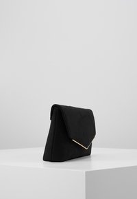 Pochette enveloppe en suède noir avec un bord en métal doré sur le rabat, reposant sur un piédestal blanc contre un fond gris clair.