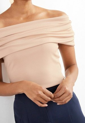 Vrouw draagt een beige off-shoulder geribd topje met een marineblauwe rok, handen voor zich geklemd, en een getextureerde ronde oorbel zichtbaar aan haar linkeroor.