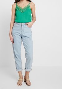 Femme portant un caraco vert orné de dentelle, un jean bleu clair retroussé, et des sandales à talons beige, debout avec une main dans la poche.