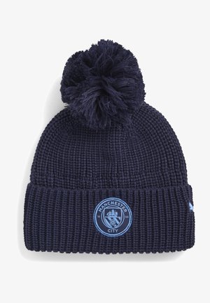 Gorro de punto azul marino con pompón, con el emblema del club de fútbol Manchester City en el puño doblado.