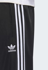 Svarta träningsbyxor med vit trestripesdesign på sidorna, med en broderad Adidas-logotyp nära midjan. Slät yta.