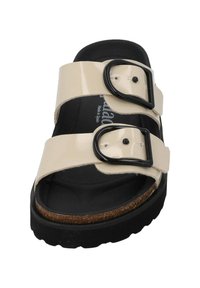Beige patentleren sandaal met twee brede banden, zwarte gespen en een EVA-zool. Beschikt over een kurken voetbed voor comfort.