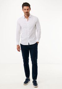 Camicia bianca con maniche lunghe, abbinata a chinos blu navy e sneakers blu navy, in piedi davanti a uno sfondo bianco semplice.