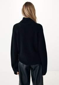 Pull en tricot noir à col roulé avec texture côtelée, épaules tombantes et manches larges, porté avec un pantalon en cuir noir sur un fond uni.