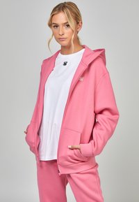Roze ritsjack in een zachte, gladde stof met een capuchon en voorkeurszakken, gecombineerd met een wit t-shirt en bijpassende roze broek.