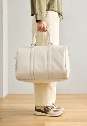 Balta odinė sportinė kuprinė su dviem tvirtais rankenomis, pasižyminti lygiu paviršiumi ir subtiliais logotipo akcentais, laikoma žmogaus, vilkinčio šviesius kelnes.