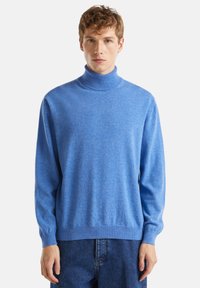 Mladý muž oblečený v světle modrém roláku a tmavě modrých džínách, stojící na jednobarevném světlém pozadí.