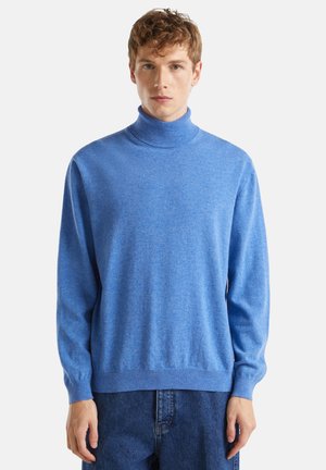 Jeune homme portant un pull col roulé bleu clair et un jean bleu foncé, se tenant contre un fond clair uniformément.