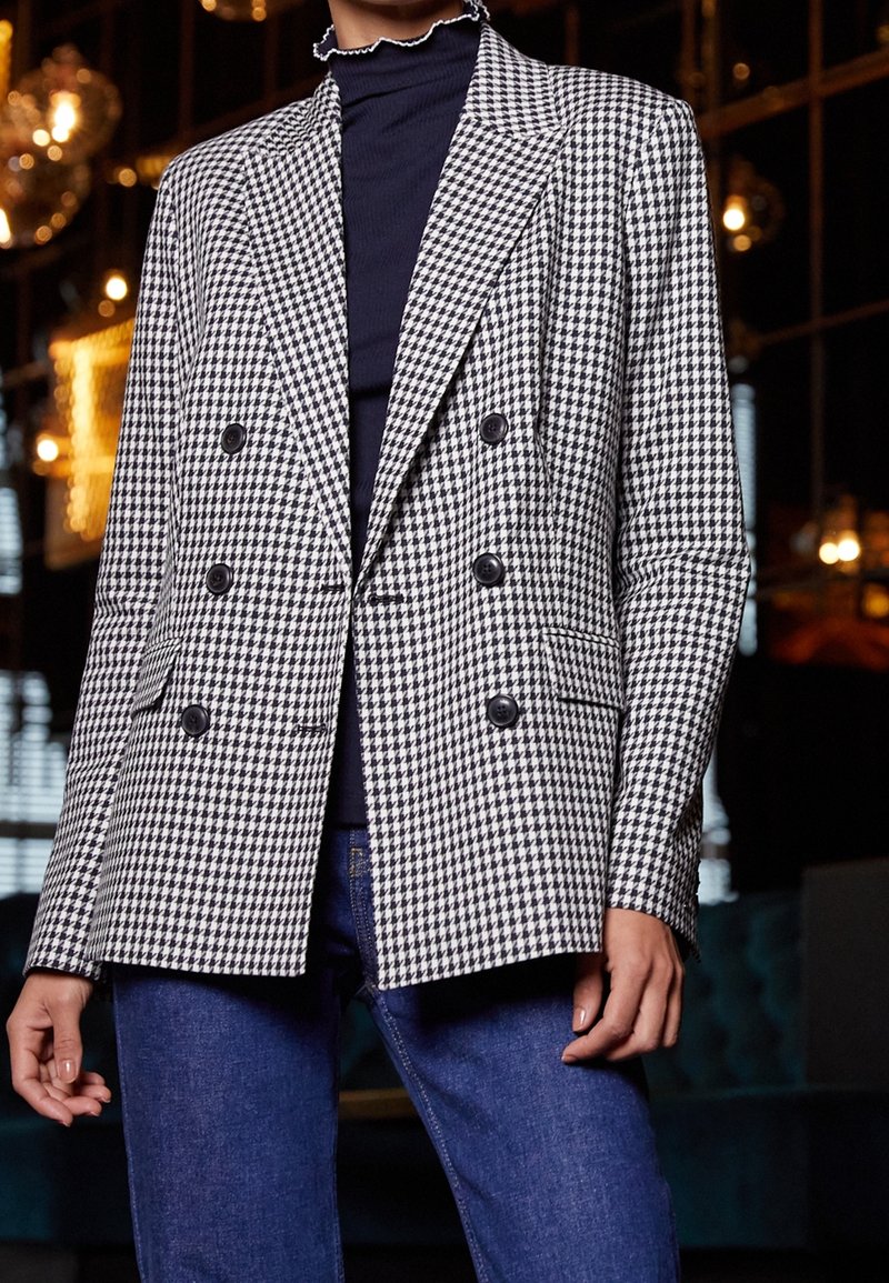 Persoon in zwart-wit houndstooth dubbelknopsgeschakeld blazer over een zwart top en blauwe spijkerbroek, staand binnen met gedimde verlichting.