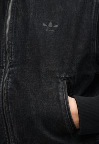 Μαύρο denim μπουφάν με φερμουάρ, λογότυπο Adidas στο στήθος και ένα χέρι μερικώς στην πλαϊνή τσέπη.