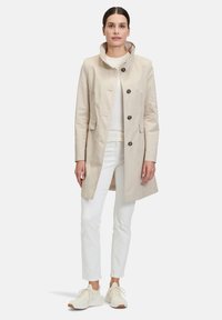 Beiger Trenchcoat mit hohem Kragen, Knopfverschluss und seitlichen Taschen, aus glattem Stoff, über einem weißen Oberteil und Hose getragen.