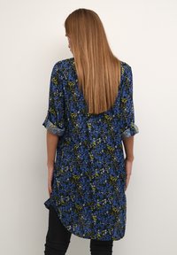 Blaue und schwarze Blumentunika mit dreiviertel Ärmeln, die grüne und weiße Akzente aufweist, aus weichem Stoff gefertigt ist und einen verlängerten hinteren Saum hat.