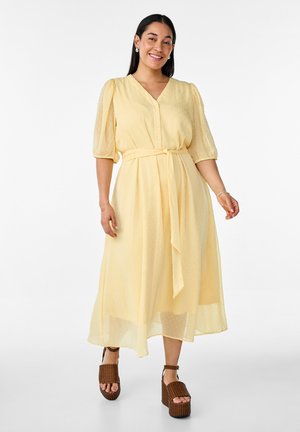 Žena sa usmieva, nosí svetložlté midi šaty s nafúknutými rukávmi a opaskom v páse, v kombinácii s hnedými sandálmi na platforme, stojaca na bielom pozadí.