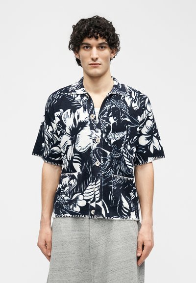 Mlad moški z kodrastimi lasmi, oblečen v črno-belo cvetlično srajco s kratkimi rokavi na gumbe in svetlo sive hlače, stoji pred belim ozadjem.