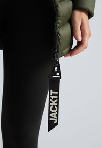 Doudoune verte olive avec fermeture éclair et une étiquette en tissu noir portant l'inscription "JACK1T." Le matériau est lisse avec une finition brillante, portée avec des leggings noirs.