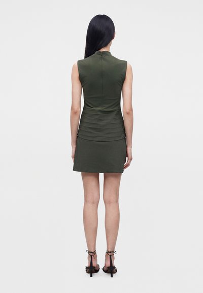 Femme aux longs cheveux foncés de dos, portant une mini-robe olive sans manches et des sandales à talons noires à brides, sur un fond uni clair.