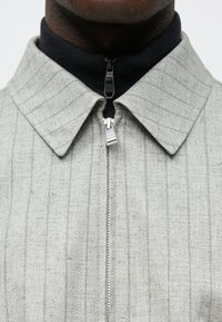 Ανοιχτό γκρι μπουφάν με υφή υφάσματος και κάθετες ρίγες. Διαθέτει κλείσιμο με φερμουάρ και πλεκτό γιακά σε ναυτικό μπλε χρώμα.