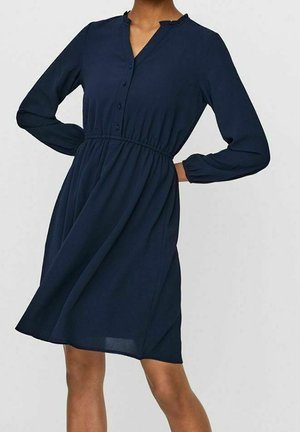 Femme portant une robe bleu marine à manches longues, longueur genou, avec des boutons sur la poitrine et une taille cintrée, posant avec une main sur la hanche.