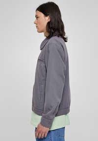 Chaqueta gris de algodón con cuello, bolsillo lateral y mangas con puños, llevada sobre una camisa verde clara y jeans azules.