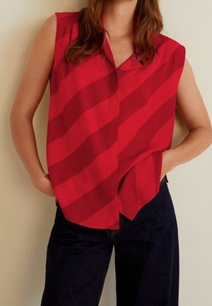 Czerwona koszulka bez rękawów z szerokimi ukośnymi paskami, włożona do ciemnoniebieskich dżinsów, ręce w kieszeniach, torso widoczne na jednolitym tle.