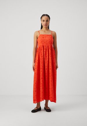 Rode maxi jurk met dunne bandjes, bloemenborduursel en een vloeiende rok. Gecombineerd met zwarte platte schoenen voor een complete look.