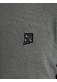Grauer gestrickter Pullover mit einem kleinen schwarzen Stoffpatch, das ein weißes geometrisches Design zeigt. Die Textur ist glatt mit gerippten Details.