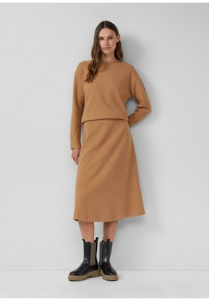 Frau trägt einen camel-farbenen Pullover mit langen Ärmeln und einen passenden Midirock, kombiniert mit schwarzen klobigen Stiefeln, und steht vor einer schlichten Wand.