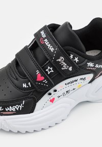Adidași alb-negru cu două benzi Velcro, text și grafice decorative, guler căptușit și talpă texturată. Detalii notabile sub formă de inimă și stea.