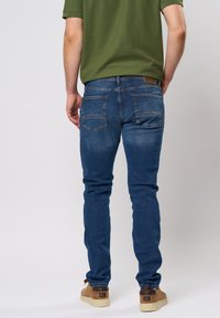 Jean denim en lavage bleu moyen, coupe slim avec un léger délavage, comportant deux poches arrière et un détail de couture moyen. Porté avec des chaussures beiges.