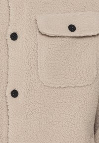Beige fleece shirt jacket with a textured surface, black buttons, and a patch pocket on the left side.