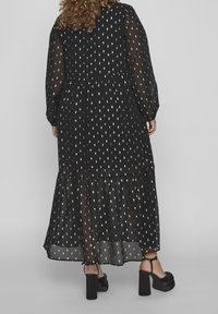 Vestido maxi negro con estampado de hojas blancas, mangas largas transparentes, dobladillo con volantes y falda en capas. Lleva sandalias negras de tacón bloque.