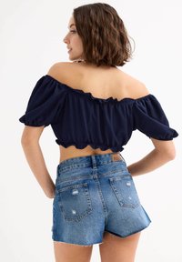 Top azul marino con escote off-the-shoulder y dobladillo con volantes, combinado con pantalones cortos de mezclilla desgastados de cintura alta con bordes deshilachados y un diseño ajustado.