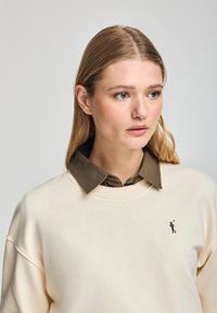 Felpa girocollo color crema chiaro con un logo sottile; sovrapposta a una camicia color verde scuro con colletto. Texture liscia, vestibilità casual.