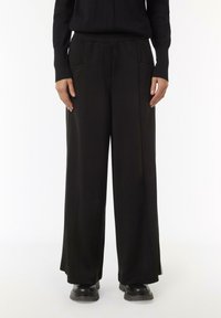 Schwarze weite Hosen mit elastischem Bund, seitlichen Taschen und glatter Textur. Kombiniert mit klobigen schwarzen Schuhen.