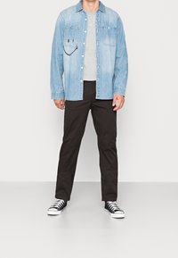 Lichtblauwe denim shirt met twee voorzakken en een smileyface-ontwerp, gecombineerd met zwarte broek en zwart-witte sneakers.