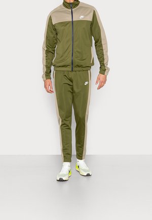 Man draagt een Nike trainingspak in olijfgroen en beige met witte en neon gele Nike sneakers, staand tegen een effen achtergrond.