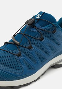 Chaussure de sport bleue en matériaux synthétiques et en maille, avec un design courbé, des lacets élastiques et une semelle en caoutchouc texturée.