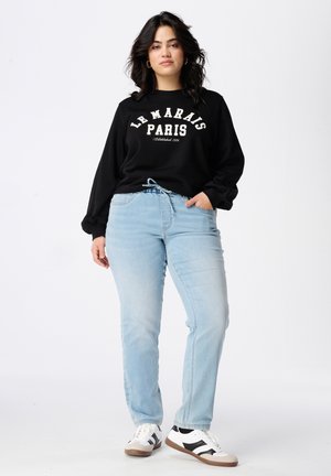 Zwarte sweatshirt met witte tekst "LE MARAIS PARIS Established 1978," gecombineerd met lichtblauwe jeans en zwart-witte sneakers.