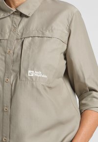 Helle beige Outdoor-Hemd aus glattem Stoff, mit durchgehendem Knopfverschluss, zwei Brusttaschen und einem Logo auf der linken Tasche.