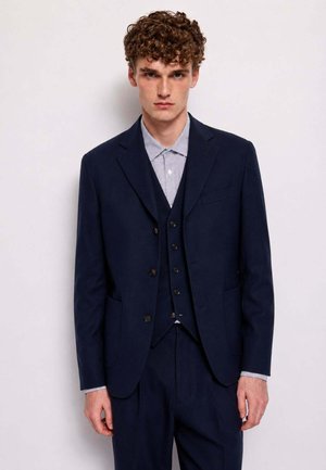 Giovane uomo con capelli ricci che indossa un completo blu scuro a tre pezzi e una camicia chiara con fantasia, su uno sfondo chiaro uniforme.