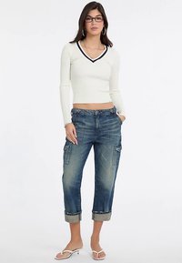 Haut à manches longues côtelé couleur crème avec un décolleté en V et un bord bleu marine, associé à un jean cargo bleu déchiré et court avec des poches.