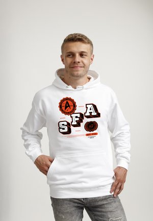 Jeune homme portant un sweat à capuche blanc avec un graphique SF Academy Starfleet noir et orange, debout, les mains dans les poches, regardant sur le côté et souriant.