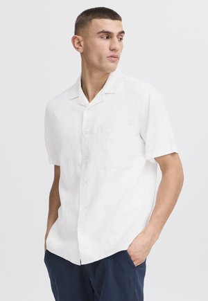 Mand iført en hvid kortærmet skjorte med knapper og brystlomme samt mørkeblå bukser, kigger til venstre med hænderne i lommerne.