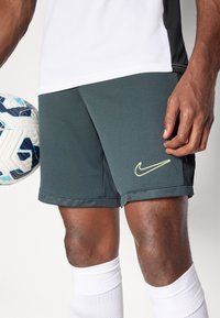 Tamnozelena Nike kratke hlače s žutim logotipom, uparene s bijelim sportskim čarapama. Hlače imaju teksturiranu površinu i opušteni kroj.