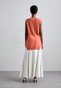 Top senza maniche di maglia arancione con texture a rete aperta abbinato a una lunga gonna bianca, che mette in mostra una silhouette fluida e un contrasto di colori.