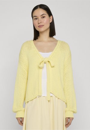 Kobieta ubrana w jasnożółty dziergany kardigan zawiązany na piersi, na białej bluzce i dopasowanej jasnożółtej spódnicy, stojąca na jednolitym tle.