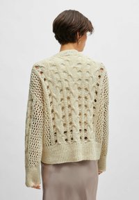 Cardigan cream lavorato a maglia con motivi a punto aperto e dettagli a trecce; presenta maniche lunghe, polsini a coste e un fondo ampio.