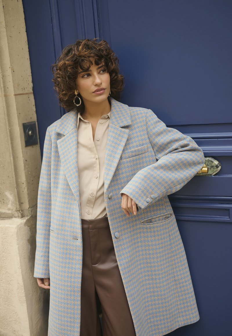 Cappotto a scacchi beige e azzurro chiaro, design sartoriale, con colletto a revers e grandi bottoni, abbinato a una blouse beige e pantaloni marroni.