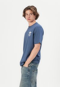 Blå bomulds t-shirt med korte ærmer og rund hals. Har hvid grafisk tekst og logo på brystet. Bæres med lyse denim jeans.